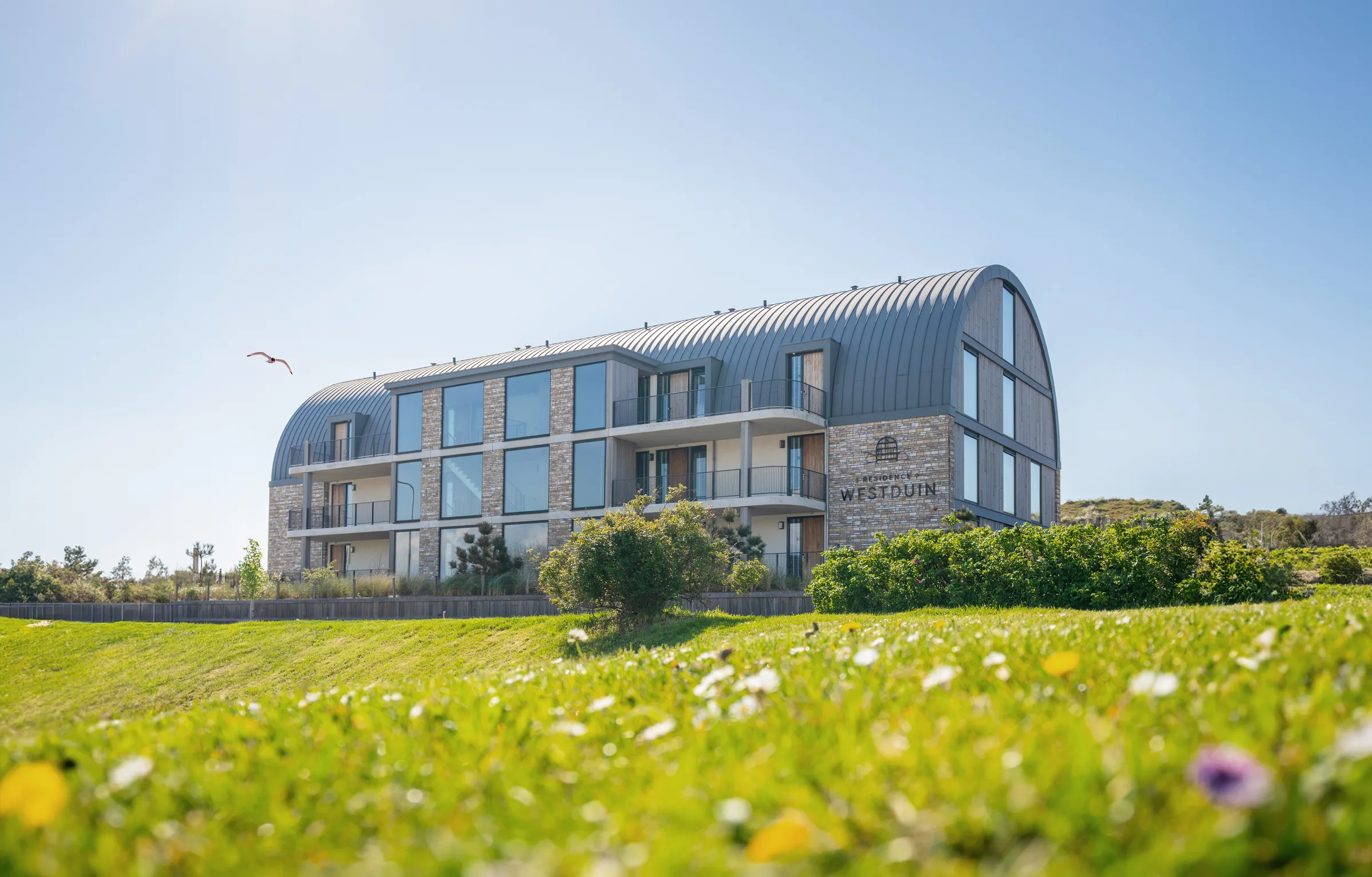 Hotel Westduin met gebogen dak achter een bloeiend duinlandschap onder een blauwe lucht