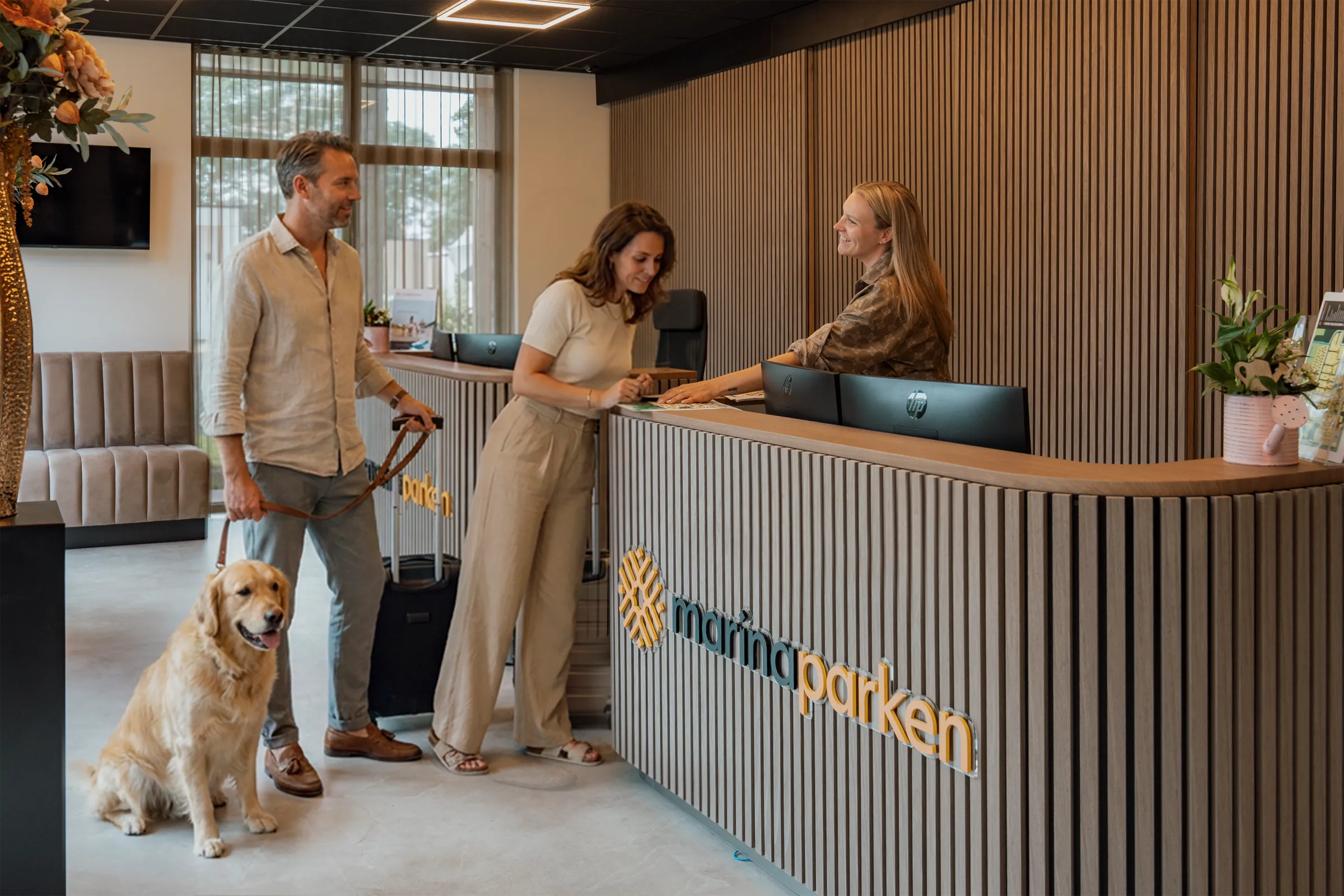 Receptiemedewerker verwelkomt een stel met koffer en hond aan de balie van MarinaParken